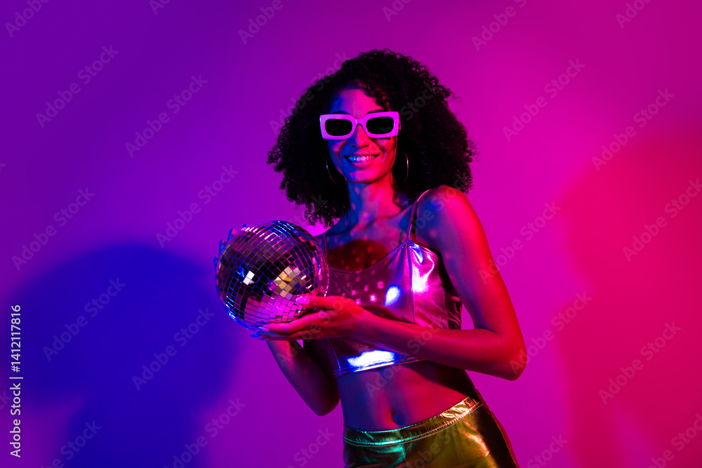 Naklejka premium Portrait of a vibrant young woman in metallic attire holding a disco ball against neon lights, evoking night club energy and retro party