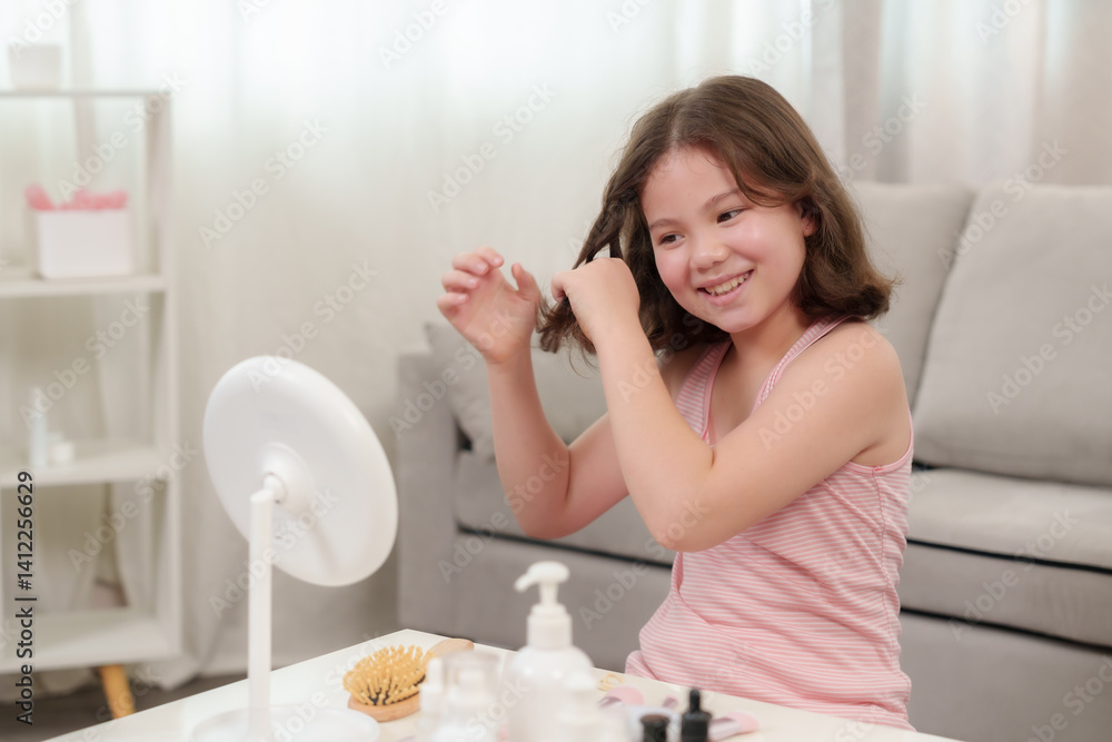 Caucasian girl wearing pink striped sleeveless top playing hair alone in living room smiling happily while sitting in front of round makeup mirror with various beauty products on white table