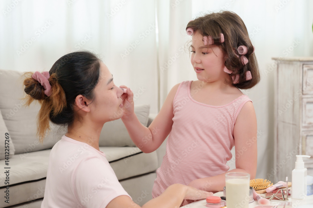 Little Caucasian girl playfully applying powder to face of Asian woman with rollers in hair while sitting together showing bonding fun connection love and shared beauty moment at home Little Caucasian girl playfully applying powder to face of Asian woman with rollers in hair while sitting together showing bonding fun connection love and shared beauty moment at home