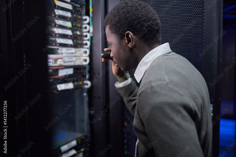 Fototapeta premium African American man in formal attire inspecting server nodes in data center. He is holding tool, examining hardware configuration, ensuring proper operation of servers