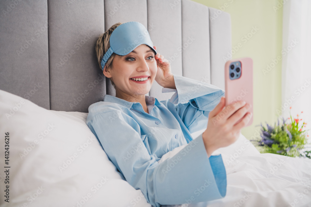 Young woman in light blue sleepwear relaxing in bed, smiling and holding smartphone during a bright morning in a cozy bedroom Young woman in light blue sleepwear relaxing in bed, smiling and holding smartphone during a bright morning in a cozy bedroom