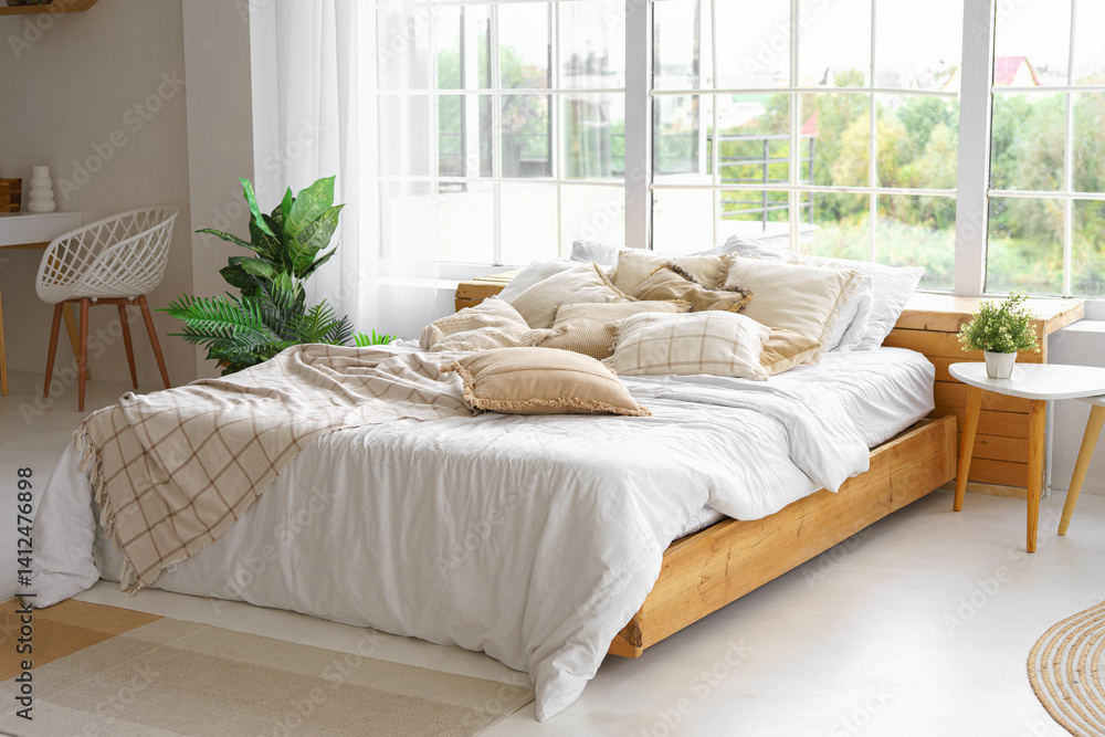 Comfortable bed with lots of soft decorative beige pillows in light sunny bedroom with green plants and large window. Interior design of living room in boho style Comfortable bed with lots of soft decorative beige pillows in light sunny bedroom with green plants and large window. Interior design of living room in boho style