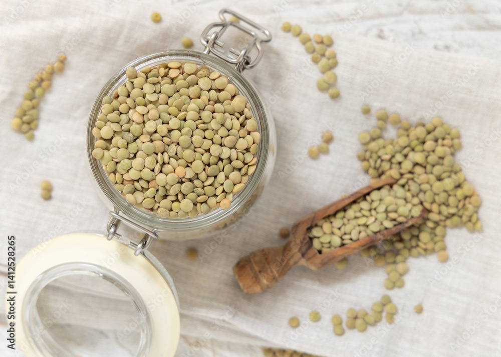 Glass jar full of raw green lentils with a wooden scoop on white table top view. Dried legumes