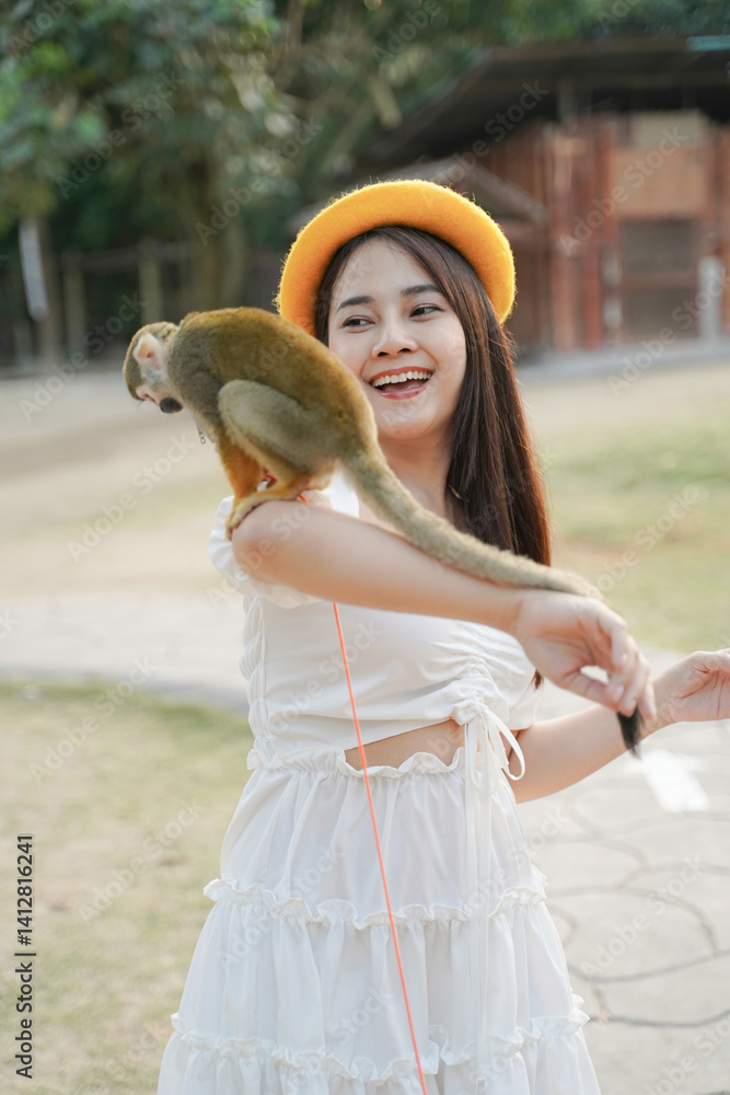 woman in white dress joyfully interacts with small monkey perched on her arm, wearing yellow hat outdoors