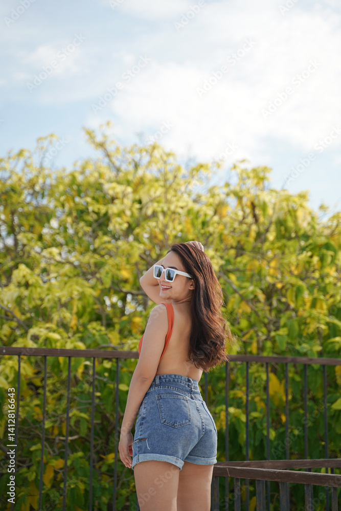 woman in sunglasses and denim shorts enjoys sunny day outdoors, standing by railing with lush greenery in background