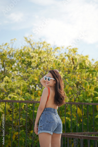 Obraz na płótnie woman in sunglasses and denim shorts enjoys sunny day outdoors, standing by railing with lush greenery in background w salonie