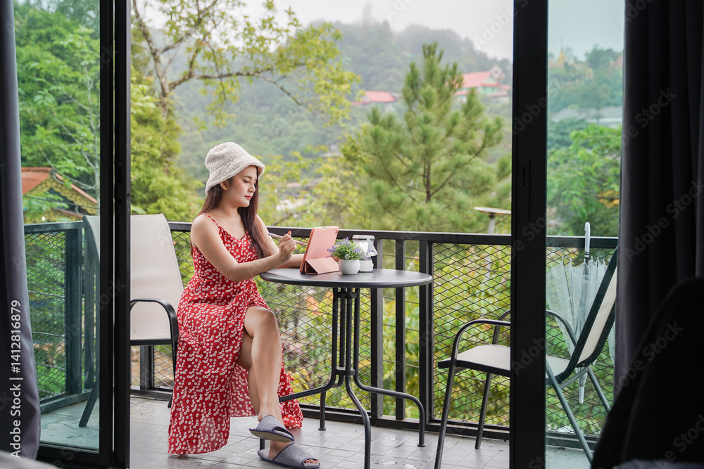 woman in red floral dress and hat sits on balcony, writing in notebook, surrounded by lush greenery