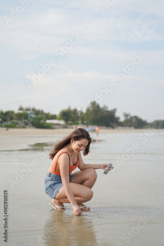 woman in bright top and denim shorts joyfully crouches on serene beach, holding sunglasses, with trees and calm water in background
