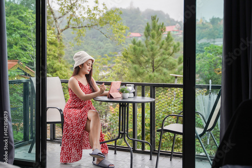 Obraz na płótnie woman in red floral dress and hat sits on balcony, writing in notebook, surrounded by lush greenery w salonie