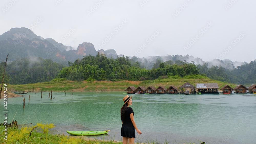 woman stands by serene lake with floating huts and misty mountains in background, creating peaceful scene