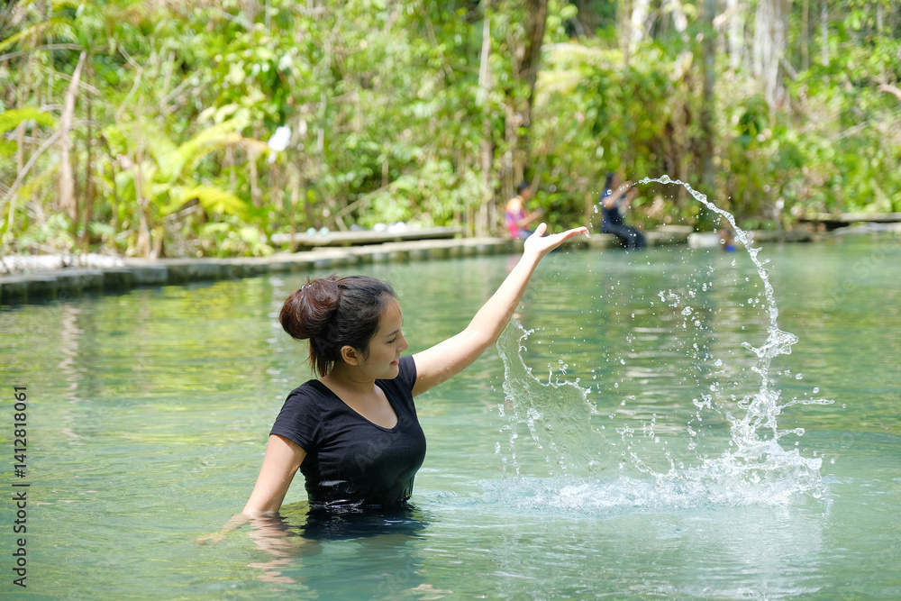 woman in black shirt joyfully splashes water in serene natural pool surrounded by lush greenery