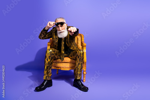 Stylish senior man in a golden patterned suit posing confidently on a purple background, showcasing elegance with a hint of eccentricity