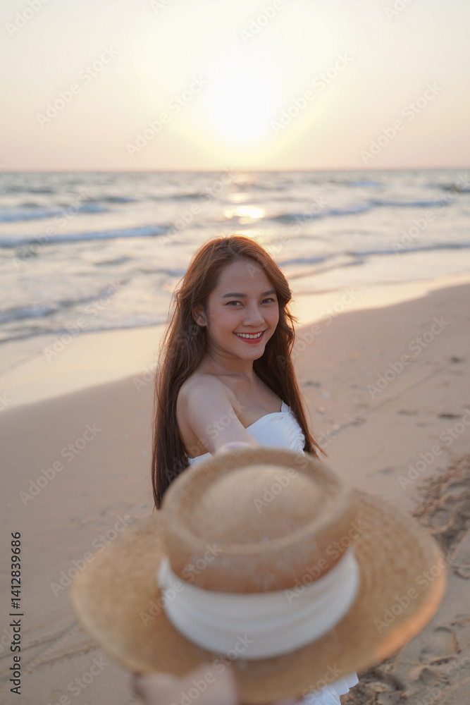 woman smiles joyfully on sandy beach at sunset, holding straw hat. ocean waves gently lap shore