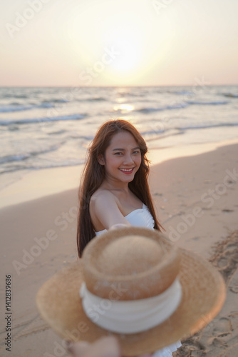 woman smiles joyfully on sandy beach at sunset, holding straw hat. ocean waves gently lap shore