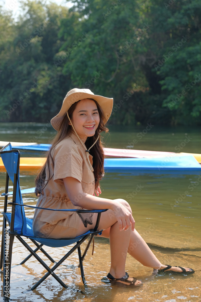 woman in sun hat sits on chair by river, smiling and enjoying outdoors
