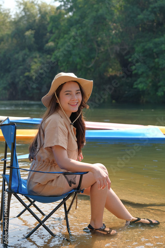 Obraz na płótnie woman in sun hat sits on chair by river, smiling and enjoying outdoors w salonie