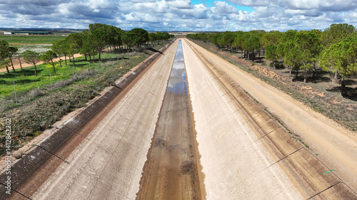 View of the Tajo-Segura water transfer canal in Albacete, Spain, with minimal water in a rural landscape with trees on both sides under a partly cloudy sky