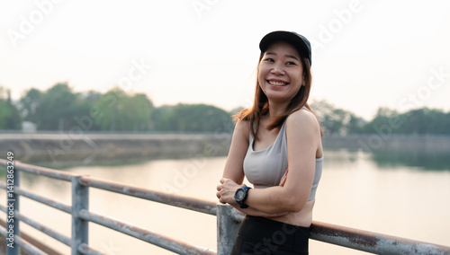 An asian woman stretches by a serene waterway, wearing a sports outfit and cap, emphasizing fitness and wellness in a natural setting.