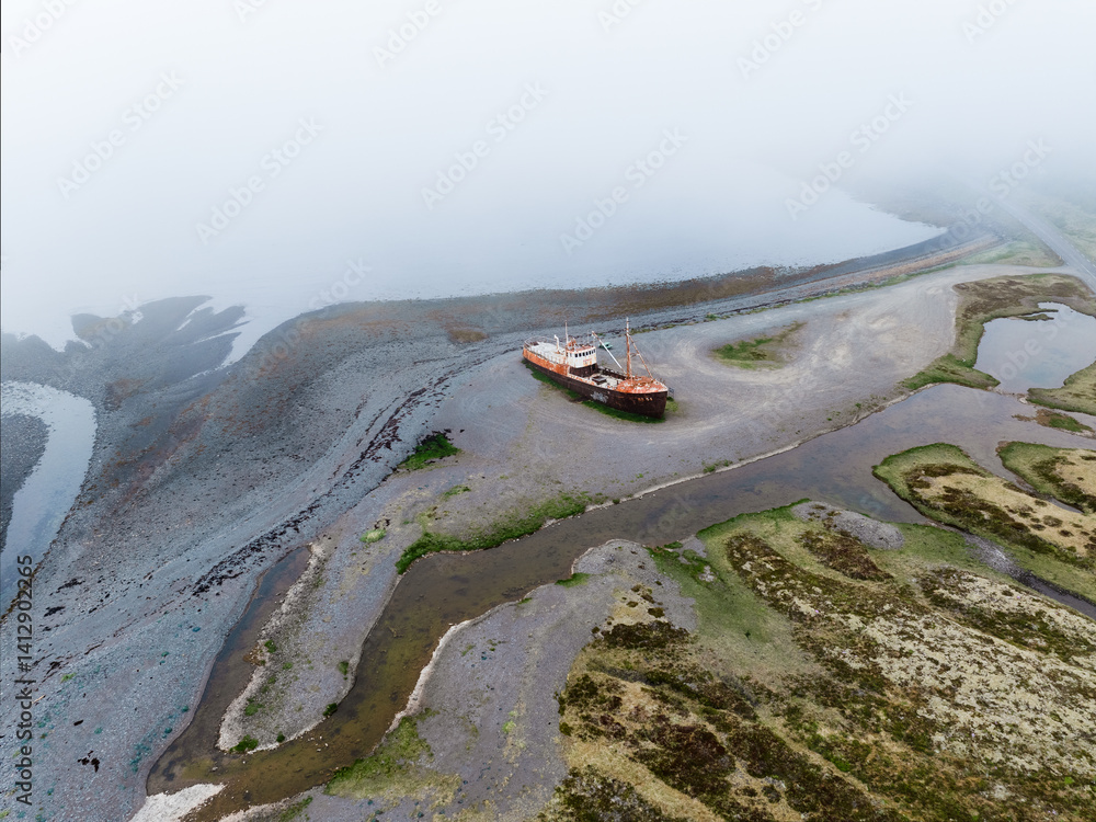 Aerial view of a rusted shipwreck stranded on a misty Icelandic shore. Surrounded by wetlands and tidal patterns, the abandoned vessel adds mystery to the remote coastal landscape.