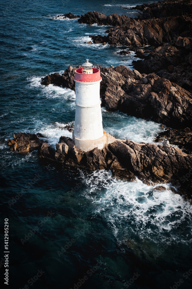 Aerial view of the lighthouse at Capo Ferro, Sardinia, standing on rugged rocks surrounded by deep blue sea and crashing waves. A dramatic coastal landmark in the Mediterranean.