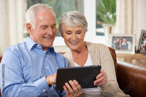 Smiling elderly couple sitting together on a sofa in the living room or outdoors, watching a tablet computer together, which they hold between themselves.