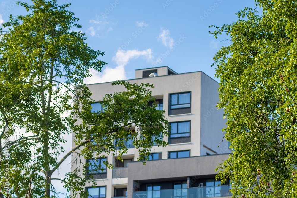 A modern apartment building in the city with a green tree in the foreground. The building is illuminated by the sun on a bright spring day, showcasing its contemporary elevation. A modern apartment building in the city with a green tree in the foreground. The building is illuminated by the sun on a bright spring day, showcasing its contemporary elevation.