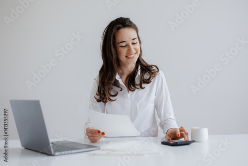 Young woman working at a desk with a laptop while using a calculator and reviewing documents in a bright office setting