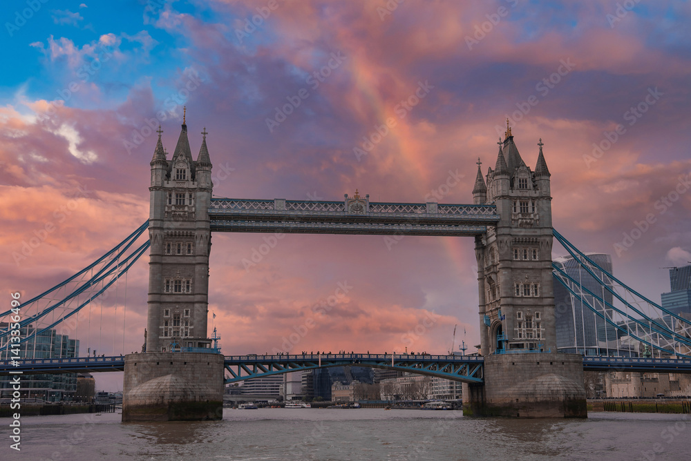 Tower Bridge spans the River Thames in London, featuring intricate towers, suspension cables, and a colorful sky. Modern buildings surround the historic landmark.