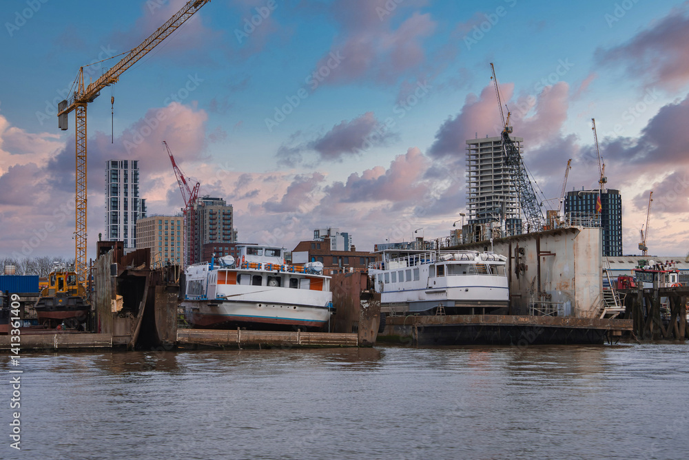 A London riverside shipyard with two white boats on platforms, surrounded by cranes and modern high rise buildings under construction at sunset.