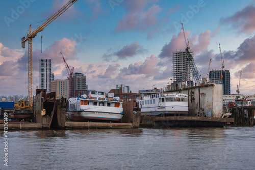Obraz na płótnie A London riverside shipyard with two white boats on platforms, surrounded by cranes and modern high rise buildings under construction at sunset. w salonie