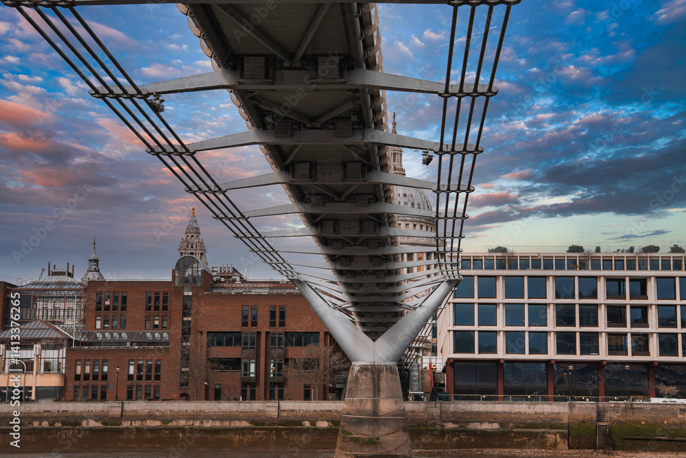 View of the Millennium Bridge's suspension cables and modern design, with St. Paul's Cathedral dome in the background under a colorful sky.