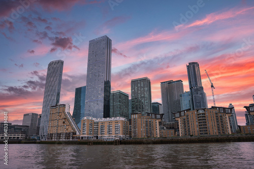 Obraz na płótnie View of London's skyline with glass skyscrapers, residential buildings, and a construction crane, reflected in calm river waters at sunset. w salonie