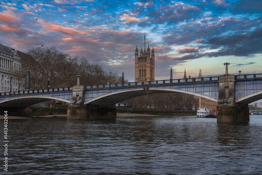 Lambeth Bridge spans the River Thames with red and white colors, framed by Victoria Tower, a boat on the water, bare trees, and a sunset sky.