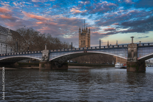 Obraz na płótnie Lambeth Bridge spans the River Thames with red and white colors, framed by Victoria Tower, a boat on the water, bare trees, and a sunset sky. w salonie