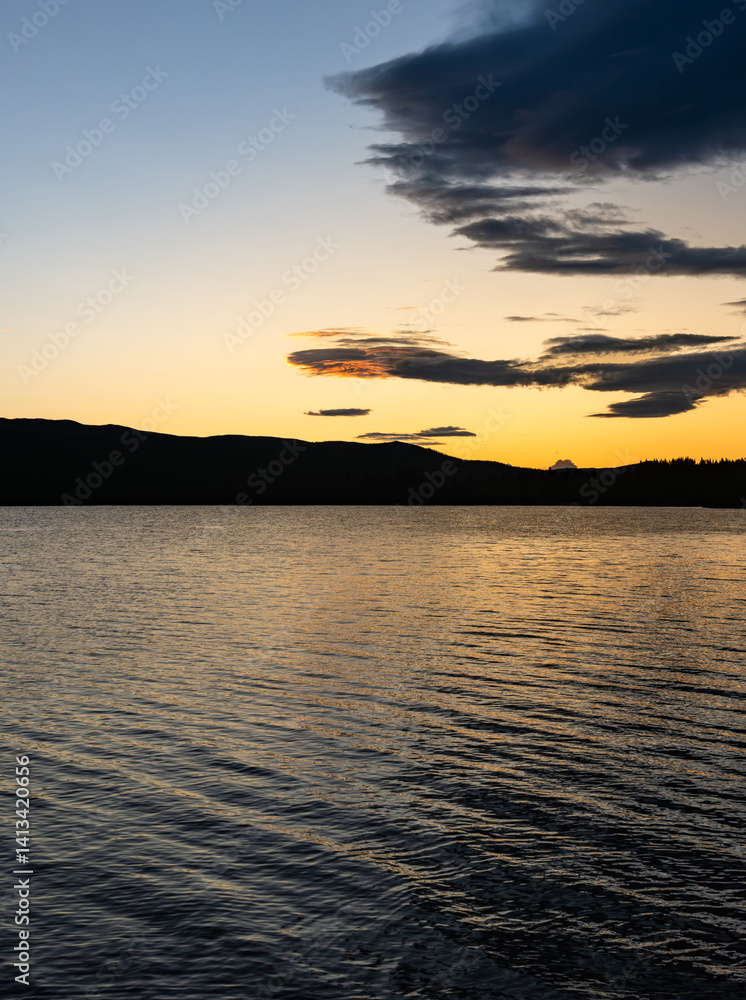Obraz premium A calm lake reflects the rich colors of sunset as the blue hour envelops the landscape. Dark silhouettes of mountains rise against the vibrant sky in Dalarna, Sweden