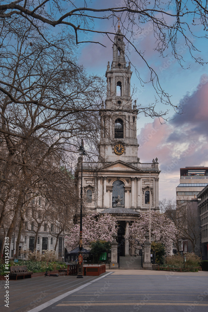 Baroque style St. Mary le Strand church in London, surrounded by trees in bloom, with a paved area, benches, and modern buildings in the background.
