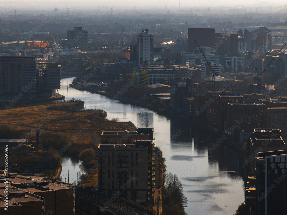 Aerial perspective of Manchester, UK, showing a river winding through skyscrapers, modern buildings, construction cranes, and patches of greenery.