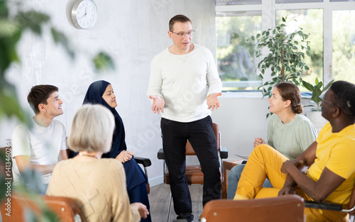Smiling friendly man conducting informal work meeting with mixed age group of colleagues of different nationalities sitting on chairs in circle in office, discussing work-related situations