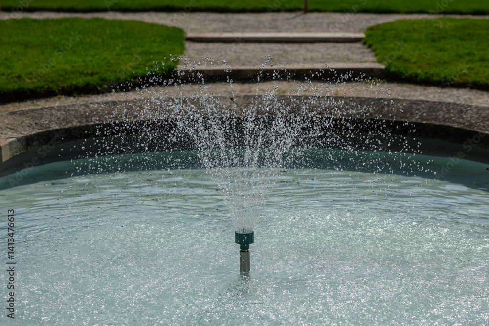 fountain spraying water in a circular pool, set against a grassy park with a stone path and trees in the background.