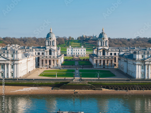 The Old Royal Naval College's twin domes flank a green space, with the Queen's House and a tree lined hill behind. The River Thames is in the foreground.