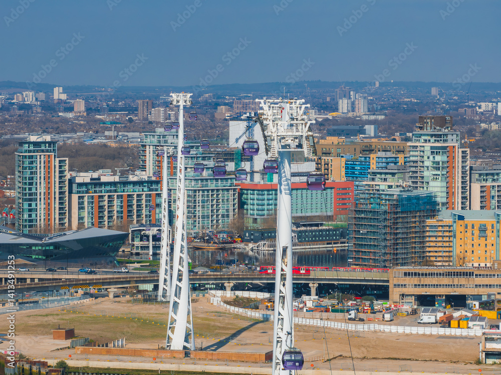 Aerial view of London featuring Emirates Air Line cable cars over the Thames, modern buildings, the O2 Arena, and urban construction areas.