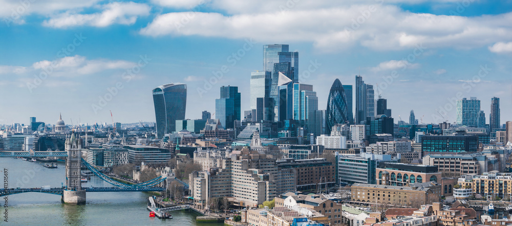 Aerial view of London featuring Tower Bridge over the River Thames, The Gherkin, The Walkie Talkie, and modern skyscrapers under a partly cloudy sky.