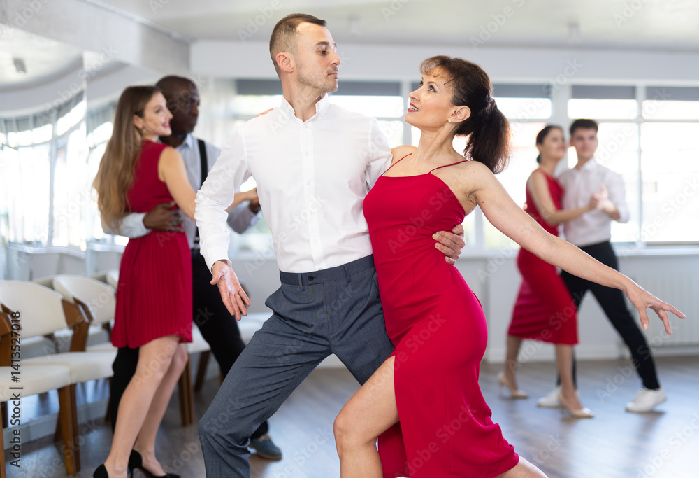 Naklejka premium Smiling young woman in red dress and her partner successfully perform mesmerizing movements of paso doble dance during dance party