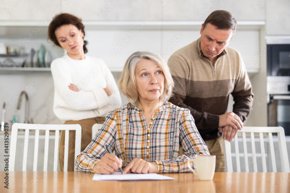 Thoughtful old woman compiling heritage document while middle-aged man and woman competing with each other in the kitchen