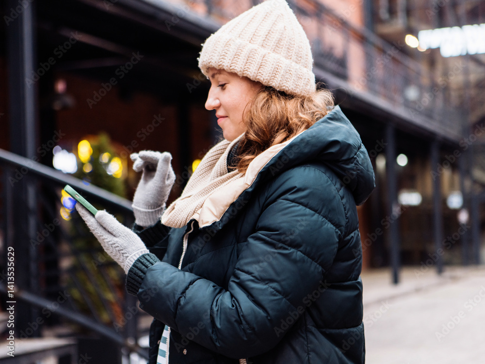 Fototapeta premium A woman in winter clothes and a hat holds a smartphone while typing a message in a winter city.