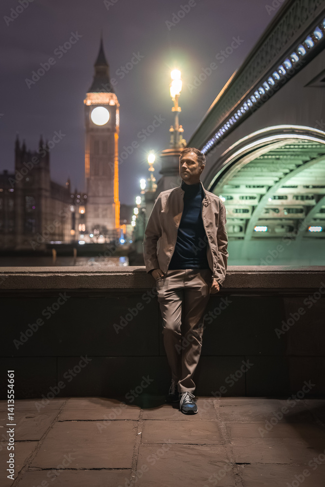 A man leans against a stone railing at night in London, with Big Ben and the Houses of Parliament illuminated. Westminster Bridge is visible to the right.