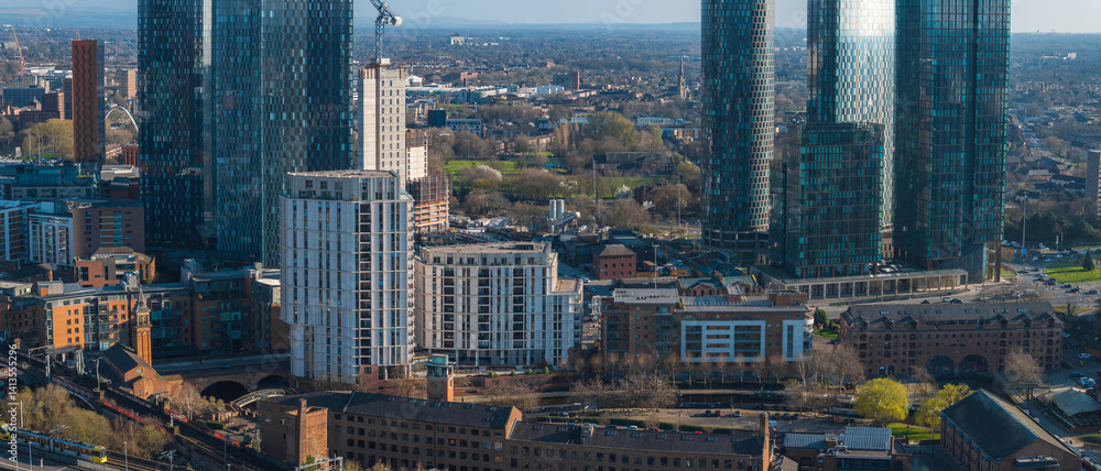 Aerial view of Manchester, UK, showing modern skyscrapers, historic brick buildings, a railway line, green spaces, and distant hills on a sunny day.