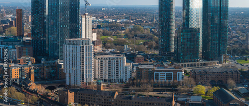 Obraz na płótnie Aerial view of Manchester, UK, showing modern skyscrapers, historic brick buildings, a railway line, green spaces, and distant hills on a sunny day. w salonie
