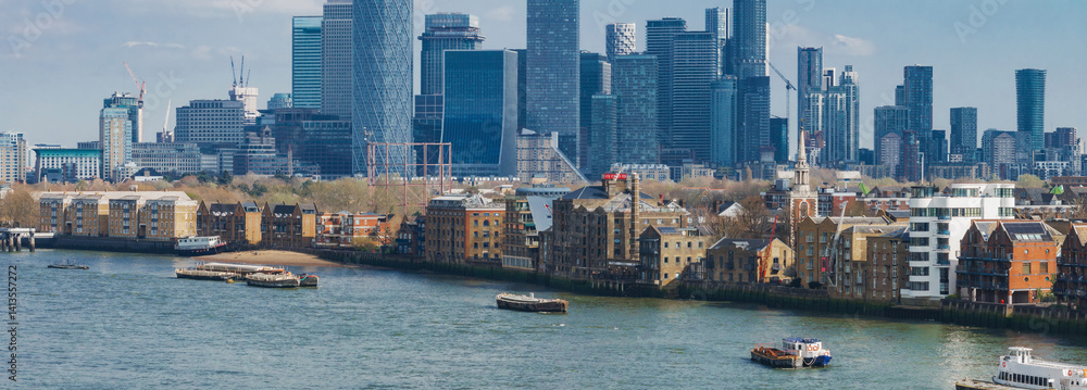 A vibrant aerial view of London's Canary Wharf skyline with modern skyscrapers, the River Thames, historic brick buildings, and boats on the water.