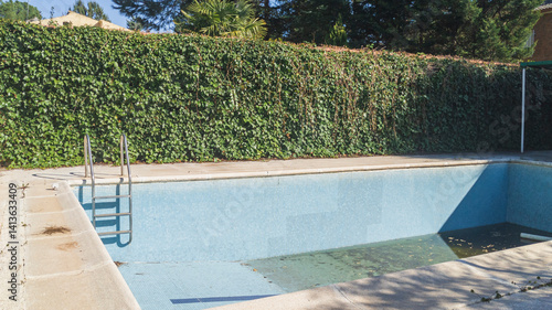 Blue tiled walls and steps of an empty swimming pool. Background wall of green leaves. Vacation low season. Dirty water with autumn leaves at the bottom of the pool